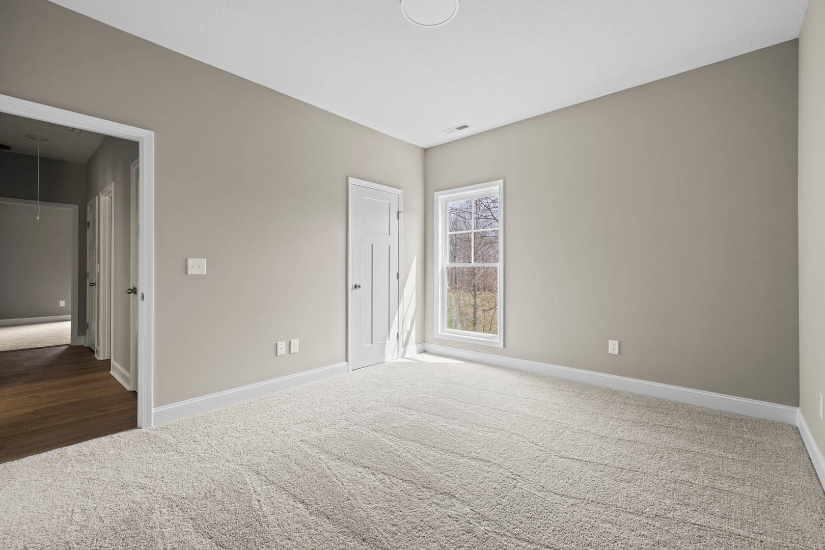 White carpeted room with a white door featuring silver knobs, window overlooking trees, wooden flooring along the edge, white ceiling with air vent, and simple molding.