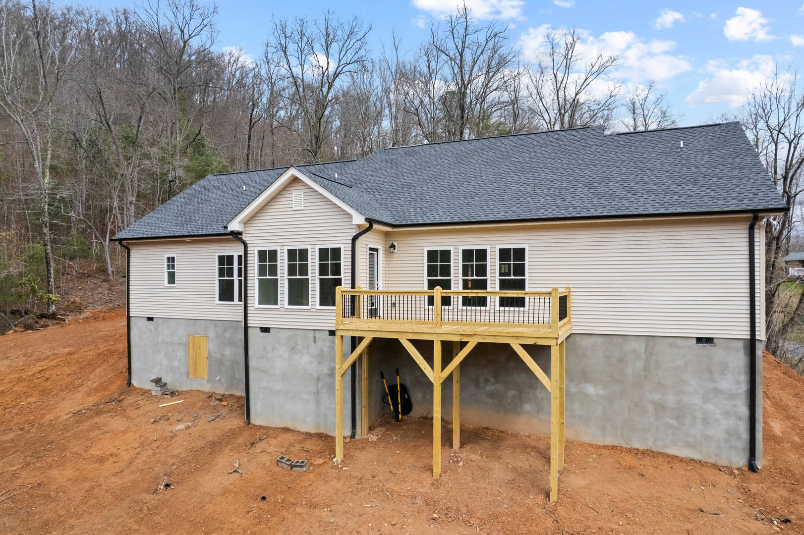 Two-story log cabin with a covered porch, elevated wooden deck, white-framed windows, and shingled roof surrounded by trees under a cloudy sky