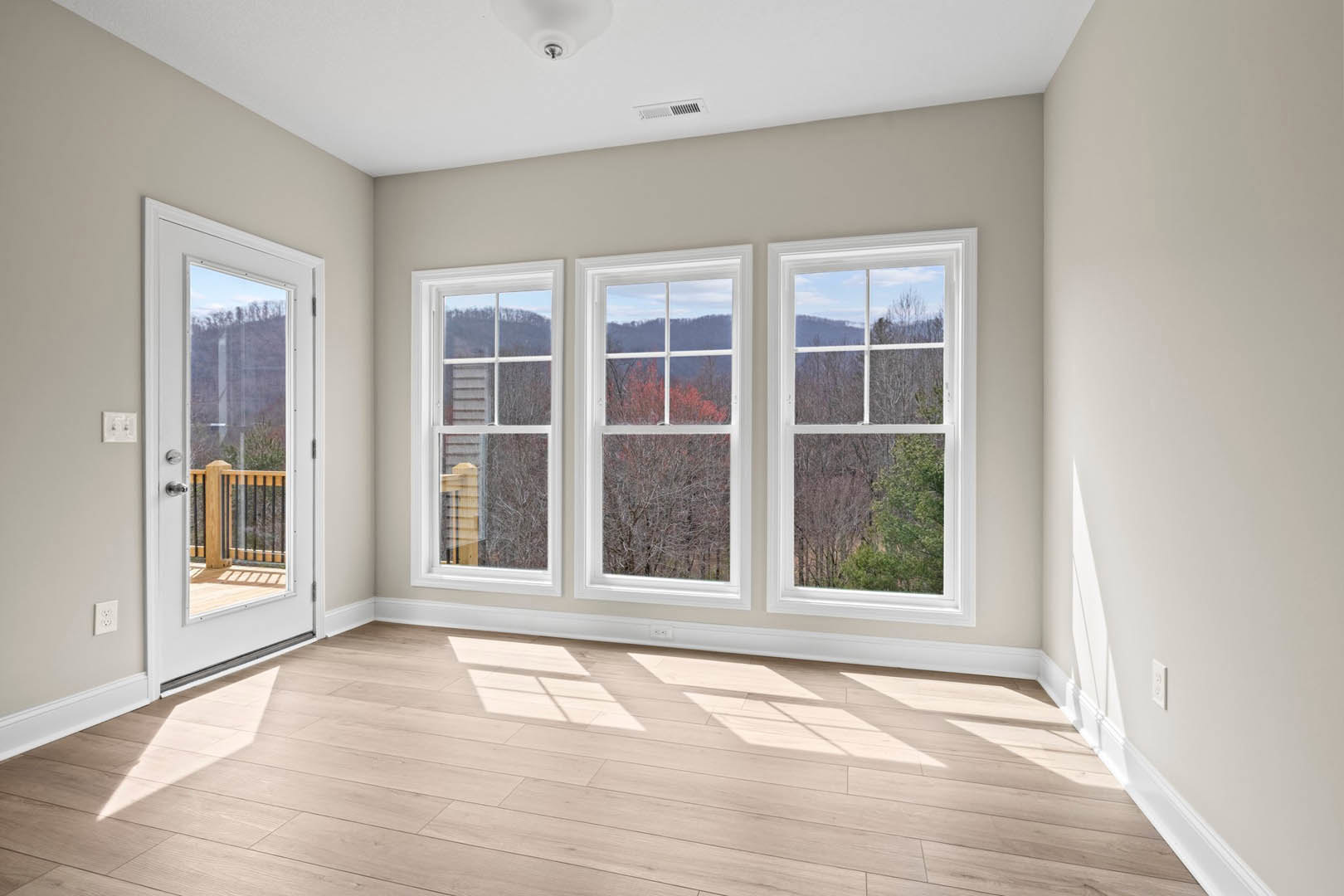 Sunlit wooden floor in a room with large windows overlooking trees and mountains, glass door opening to a balcony, white walls with a light switch
