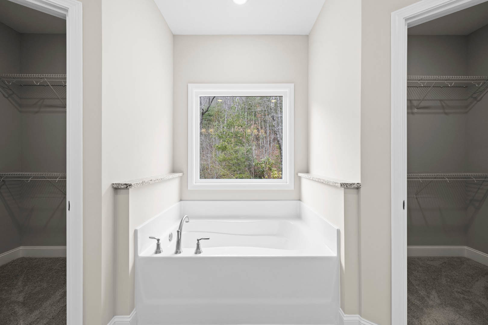 Freestanding white bathtub with chrome faucet beside a large window overlooking trees, silver bell on tub ledge, white plaster walls, recessed ceiling light, and light-colored tile