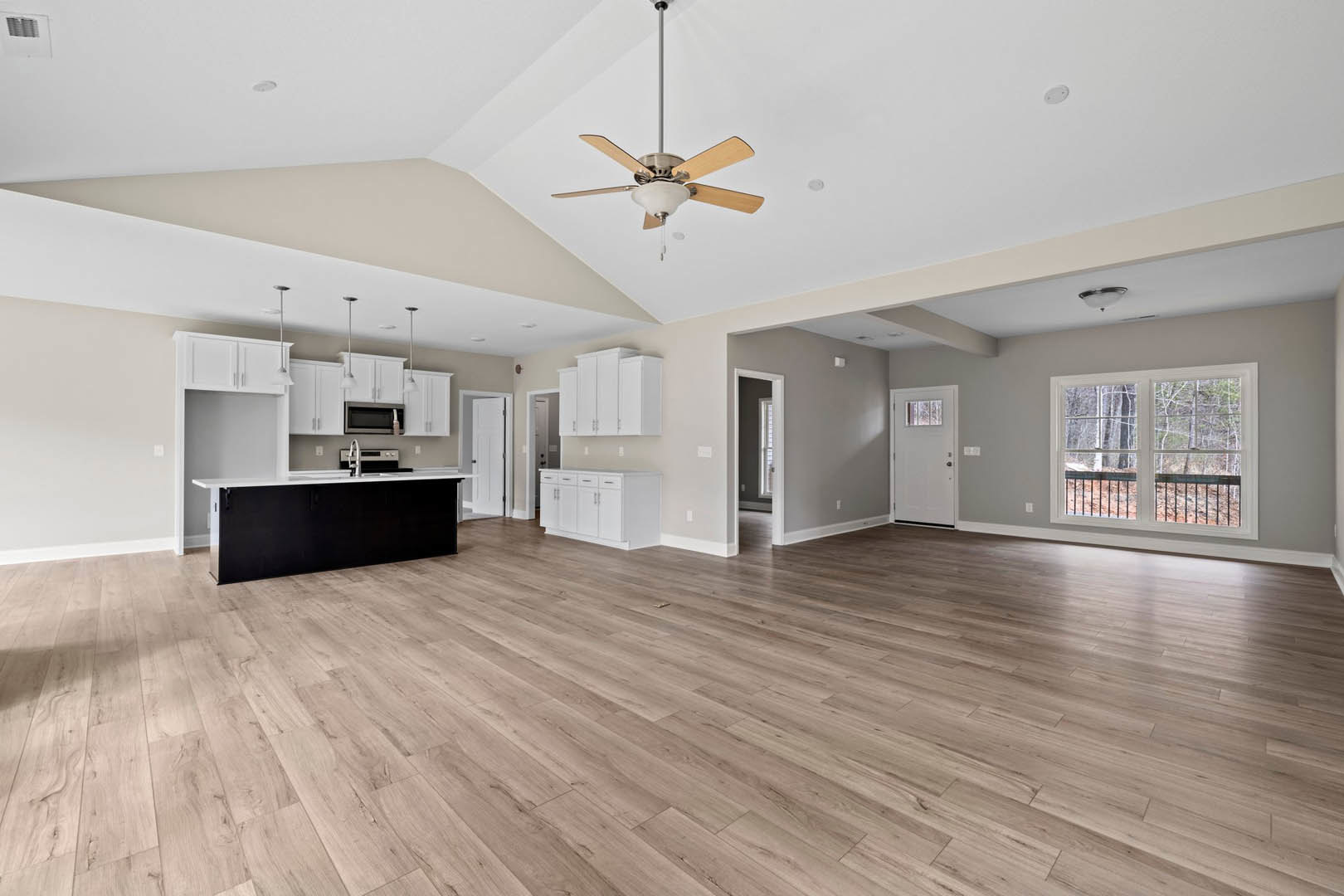 Spacious open floor plan featuring hardwood flooring, kitchen with black cabinets and white countertops, adjacent dining area, ceiling fan with light fixture, large window with