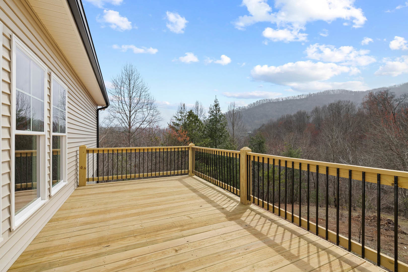 Wooden deck with black metal railing, large windows, and forested mountain view under blue sky with scattered clouds