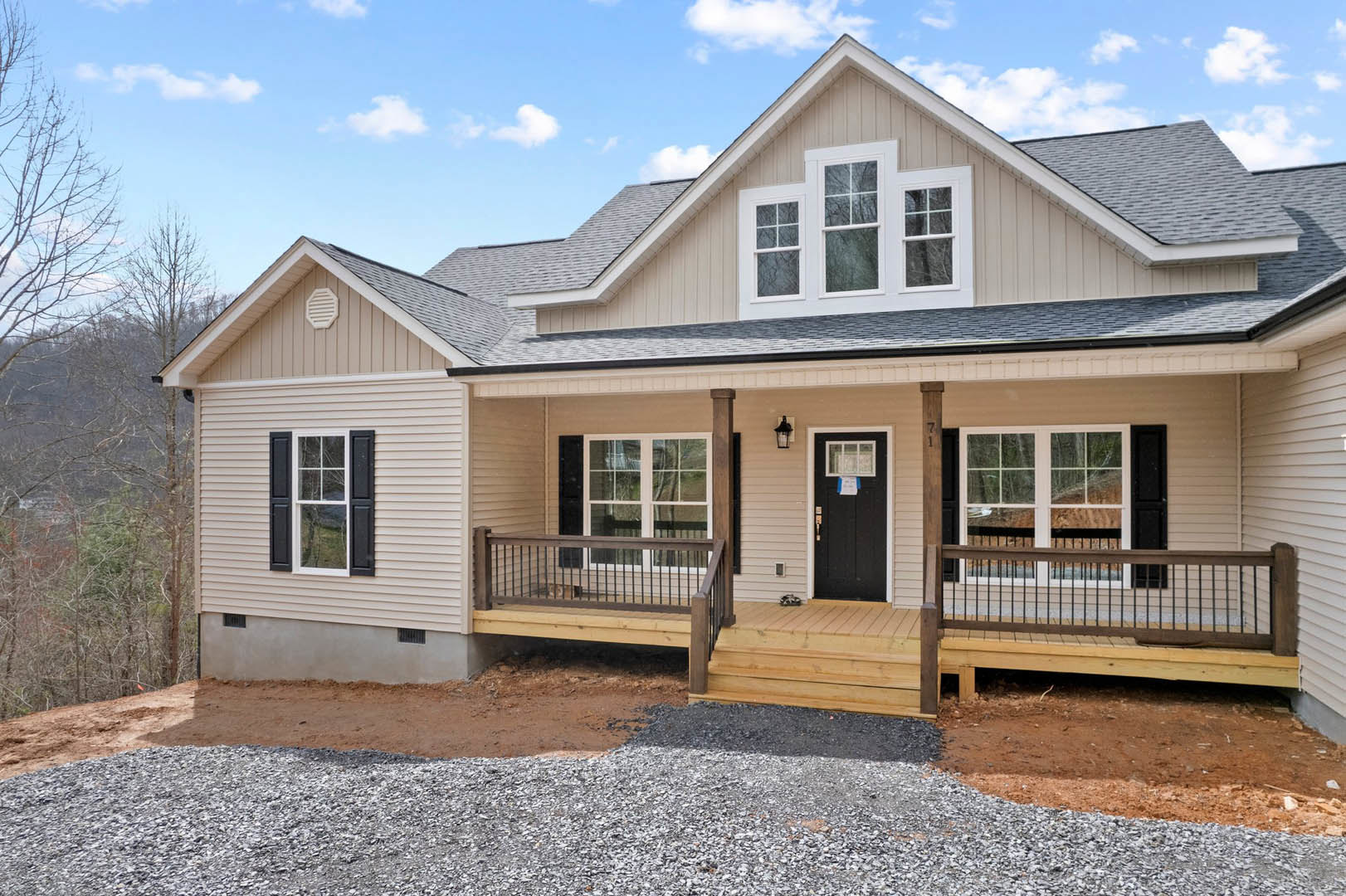 Two-story cottage with light siding, black front door featuring a white sign, white-framed windows, covered porch with metal railing, cat sitting on porch, gravel driveway in