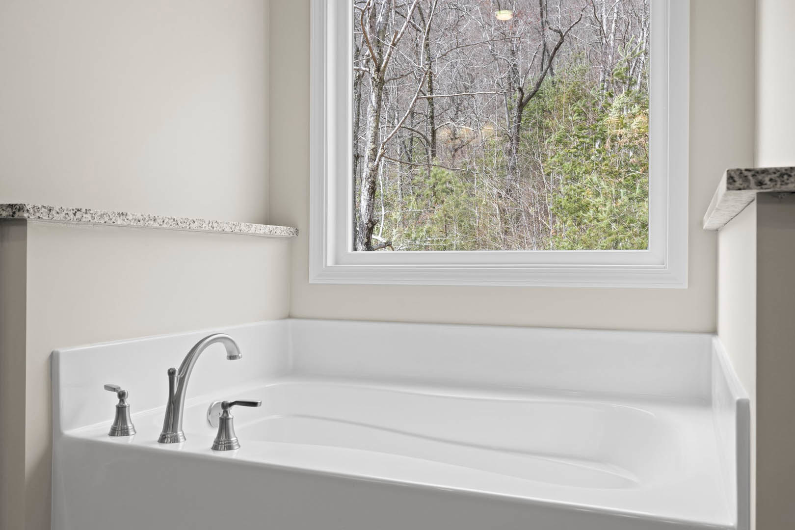 White freestanding bathtub beneath a large window with views of green trees, silver faucet and handle, light-colored bathroom walls