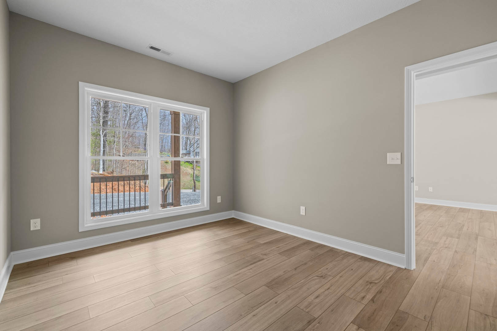 Wood flooring in a room with white plaster walls, a window overlooking trees and a road, a door, and a light switch on the wall