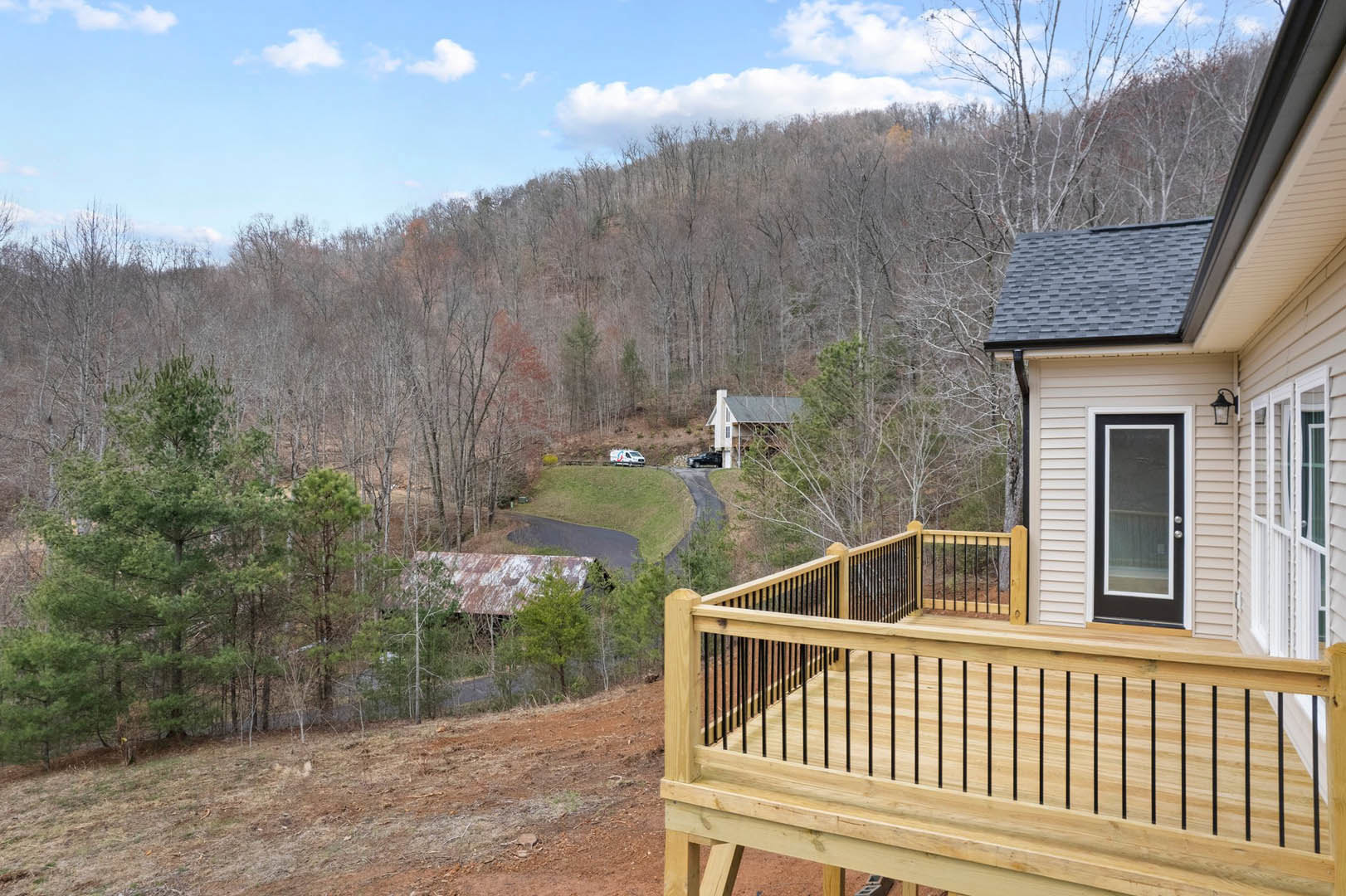 Wooden deck with black railing overlooking a house with rusty roof, surrounded by trees under a blue sky with scattered clouds