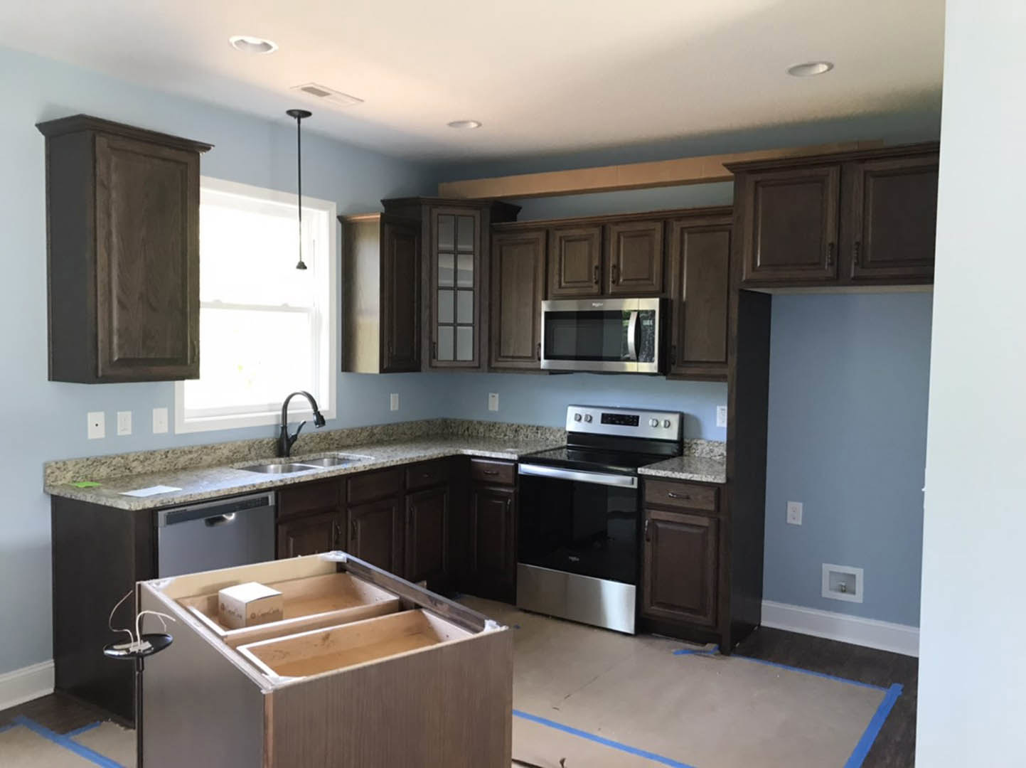 Kitchen with dark wood cabinets, light stone countertop, stainless steel microwave and dishwasher, drawer partially open with box inside, white square sink with modern faucet