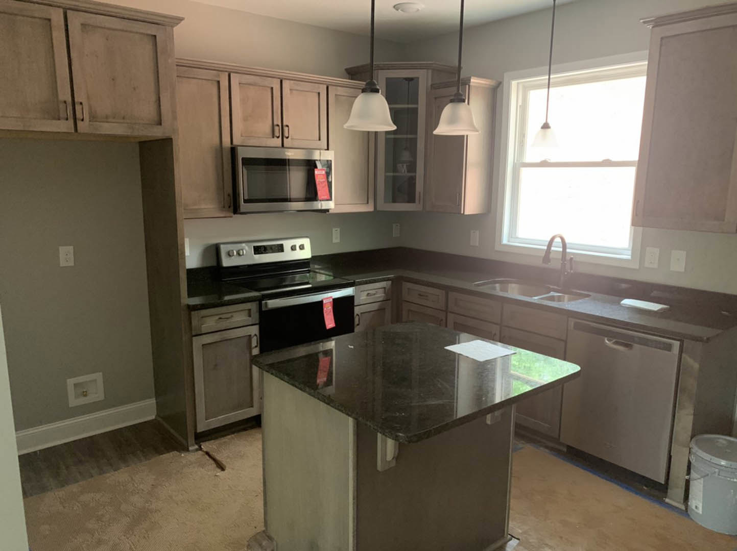 Modern kitchen with black countertop, white cabinetry, stainless steel microwave with red tag, paper on counter, bucket nearby, sunlight streaming through window, white square