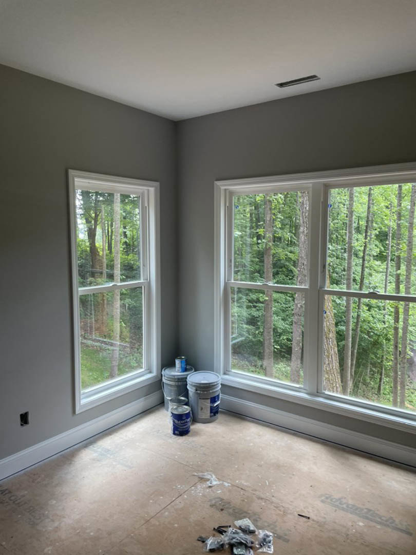 Unfinished room with two windows, paint cans and buckets on bare floor, plastic sheeting piled near wall, white bucket with blue lid, trees visible outside