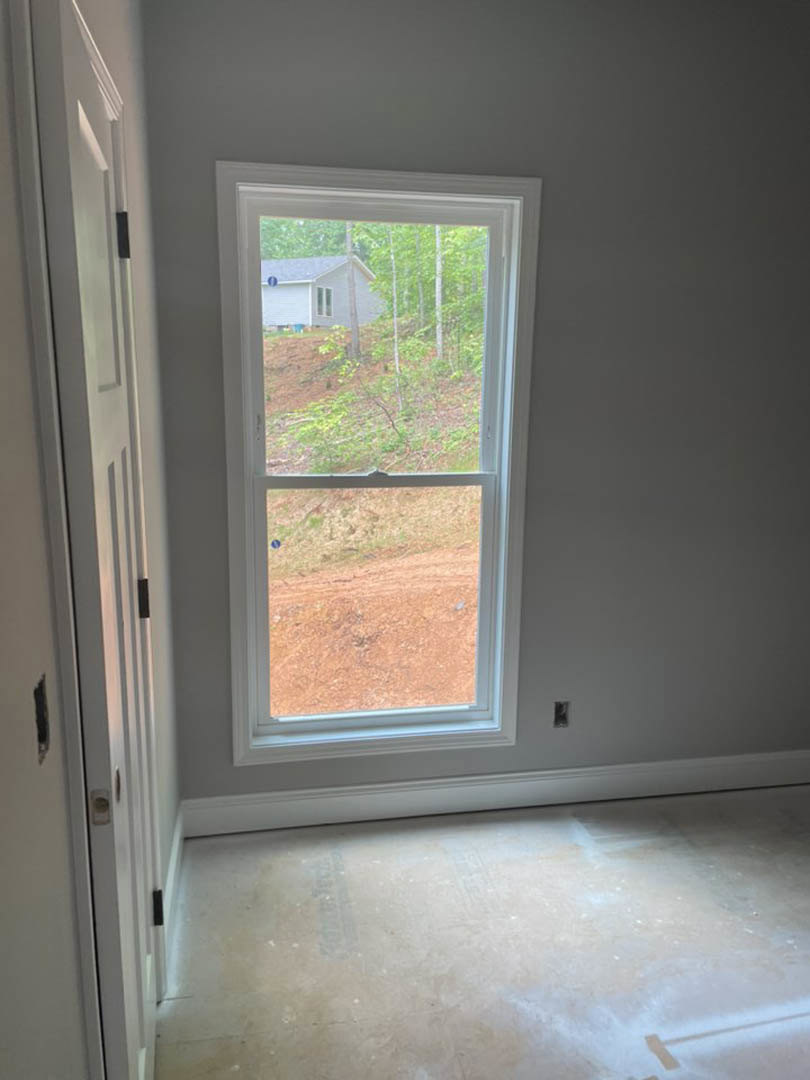 Large window set in a plaster wall, overlooking a wooded landscape with dirt and another house visible outside; light wood flooring and door frame in foreground.