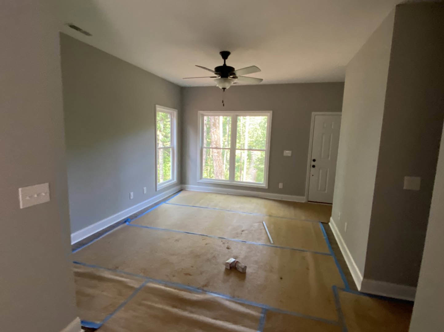 Bedroom with laminate wood flooring, two ceiling fans with lights, white door featuring black knobs, window overlooking trees, blue painter’s tape on floor marking areas.