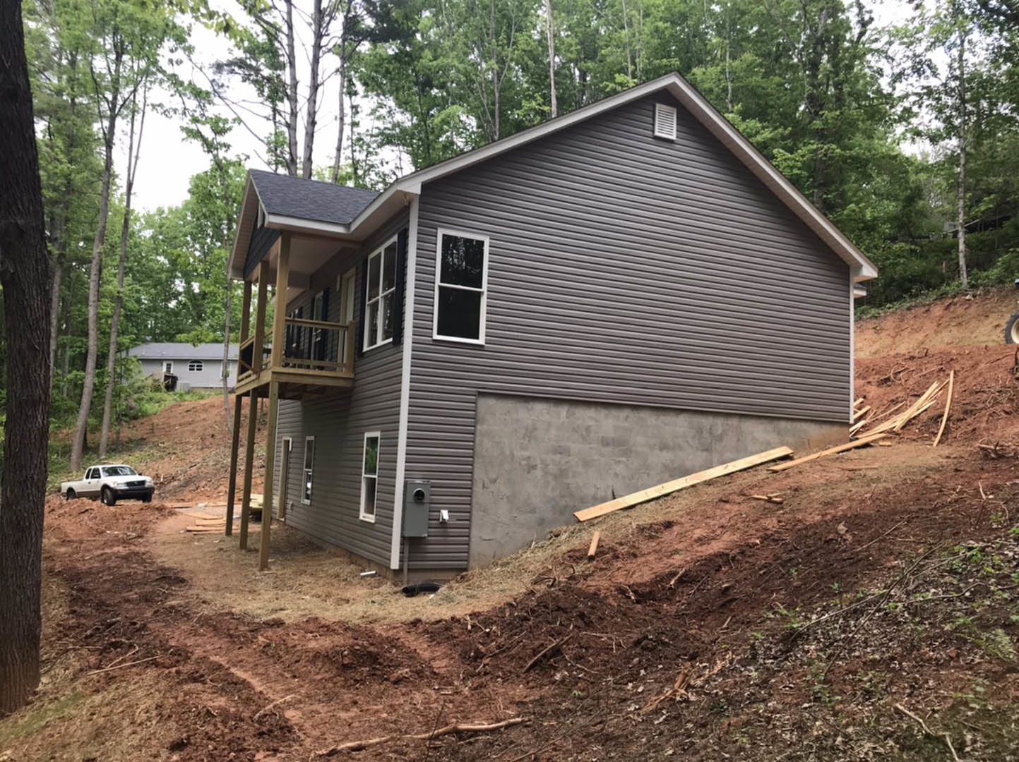 Partially built house with exposed wood framing and white window frames, situated on a dirt hill with scattered wood planks, white truck parked on unpaved road, garage attached