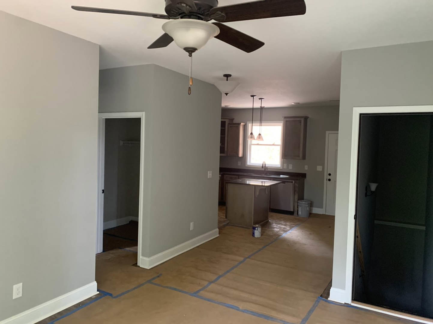 Open-concept kitchen and living area with a ceiling fan and light fixture, white walls, wood flooring, wall-mounted light, window with natural light, grey trash can, and small