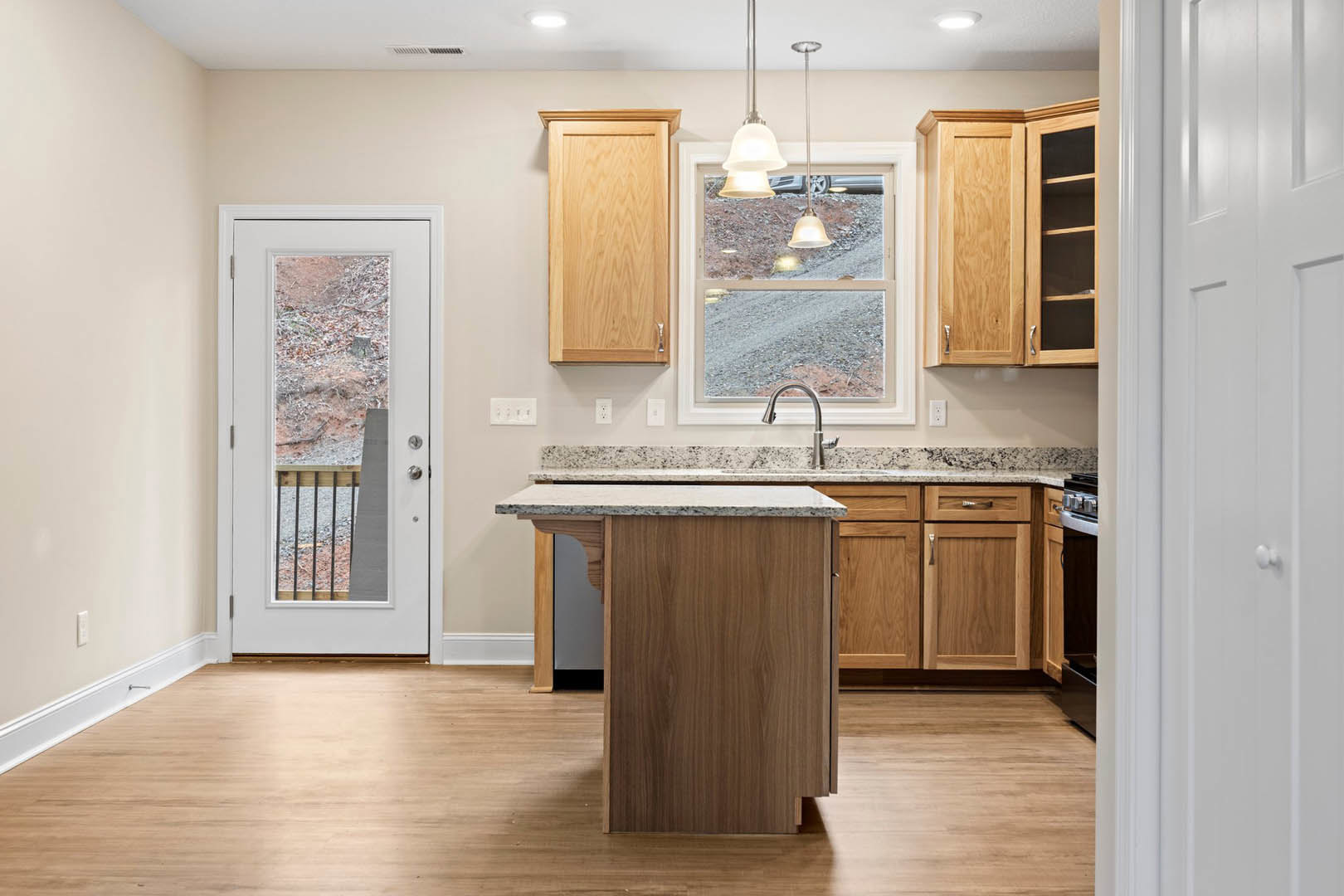 Modern kitchen featuring a central island with white quartz countertop, shaker-style cabinets, stainless steel sink, glass-paneled door, wide window overlooking street and parked