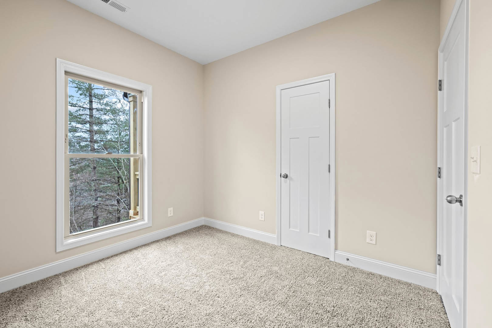 White-walled room with a white door featuring a silver knob, large window overlooking trees, light carpet in the corner, and white ceiling with simple molding.