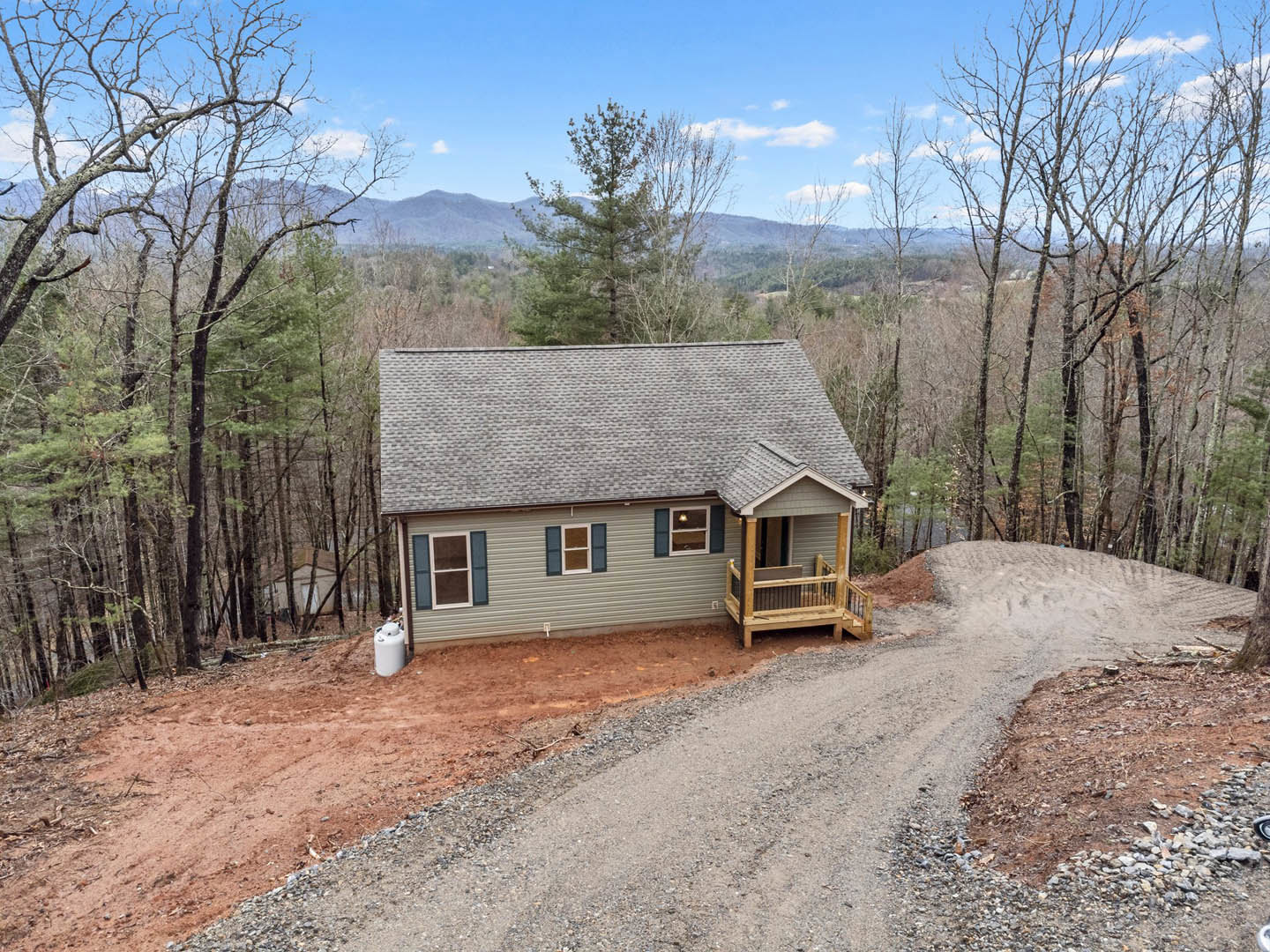 Wood-clad house with covered porch and railing, set along a gravel road, surrounded by dense forest and distant mountain backdrop