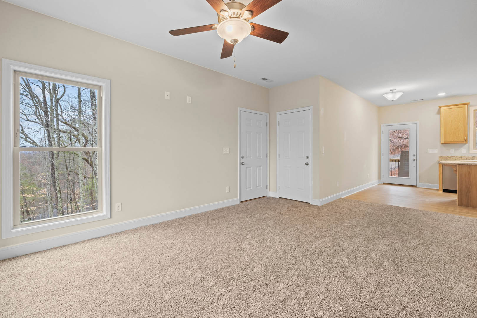 Living room with laminate wood flooring, white walls, ceiling fan with light, white door with silver knobs, large window showing trees outside