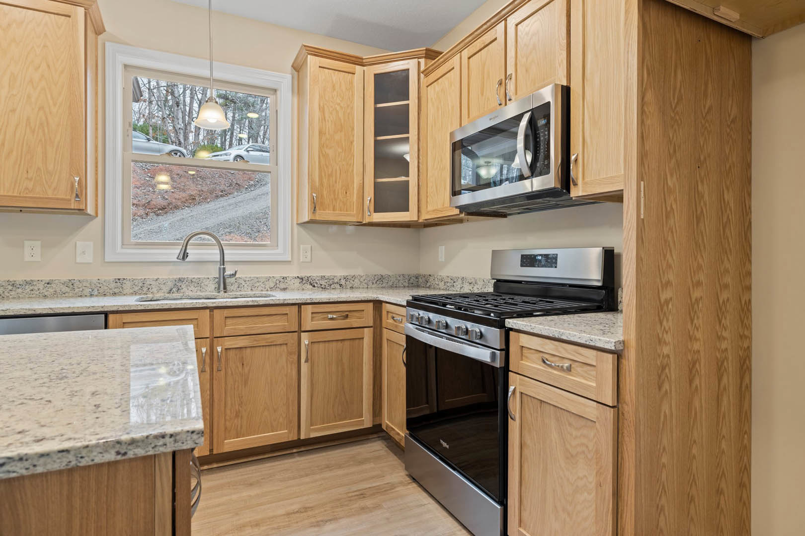 Kitchen with medium-tone wooden cabinets, stainless steel stove and oven, built-in microwave with glass door, white speckled countertop, window overlooking a road, and open
