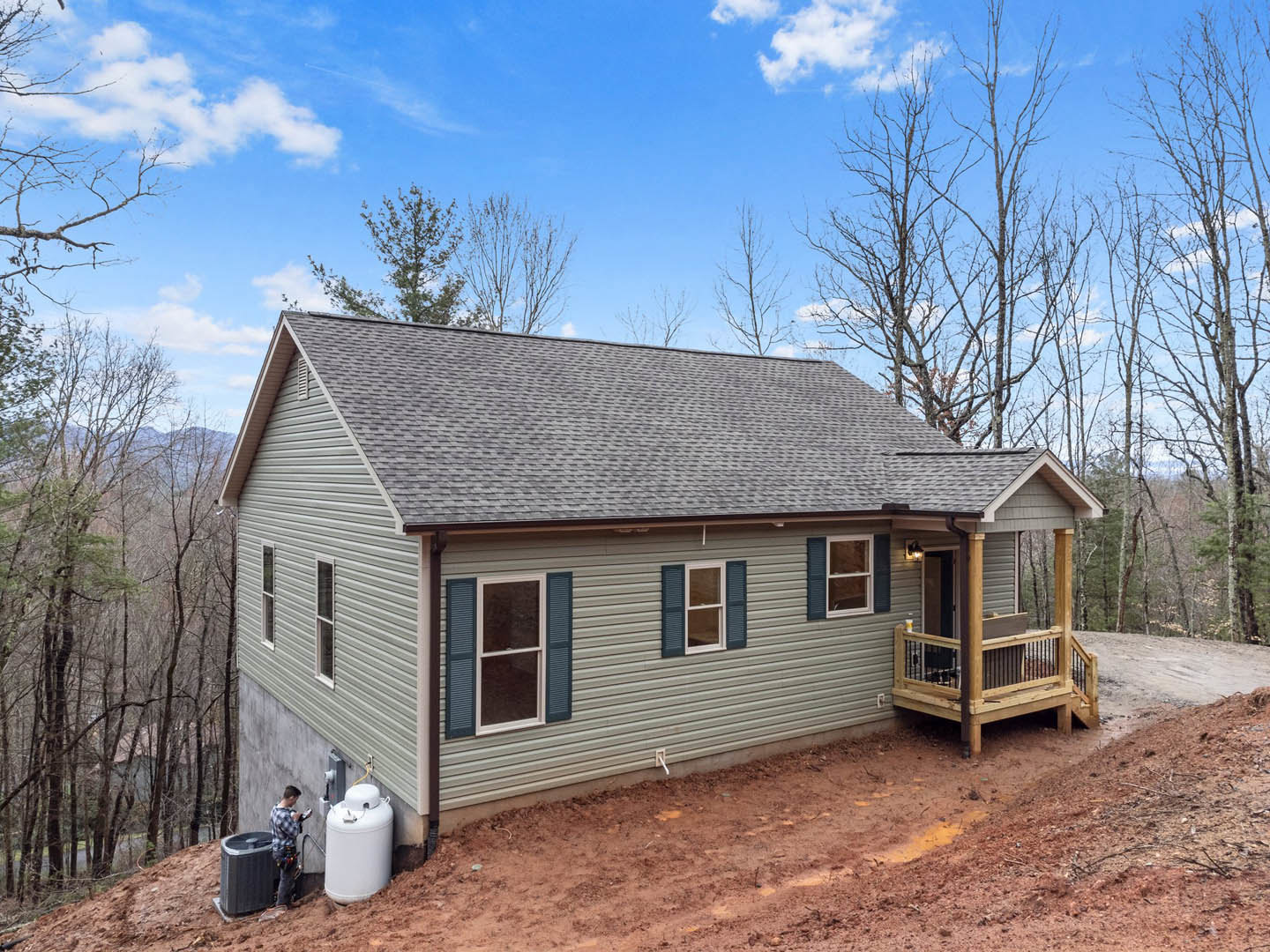 Two-story house with wood deck and gas tank on grassy hill, surrounded by trees, Wyckoff House visible in background, window with shutters on side, person in jacket near white