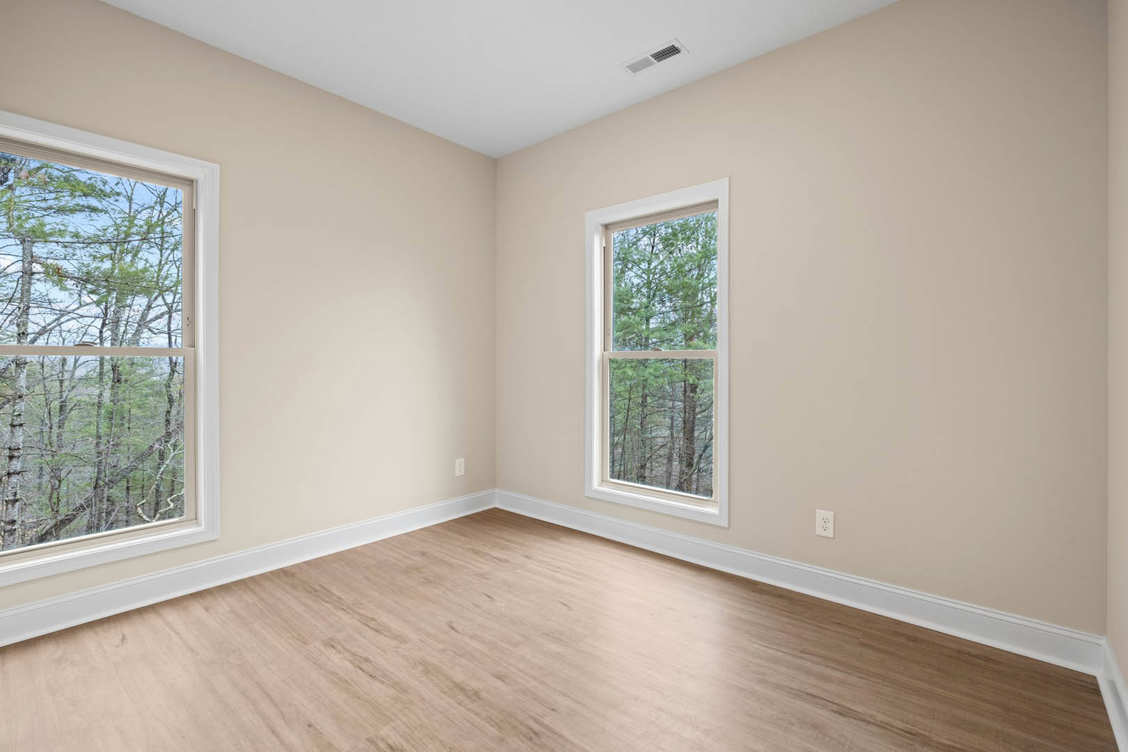 Sunlit room featuring a large window overlooking leafy trees, white plaster walls, white ceiling with recessed vent, and warm wood laminate flooring.