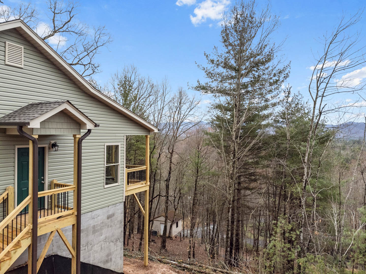 Two-story home with white siding, green front door, wooden porch and balcony, white-framed windows, metal railing, surrounded by mature trees.