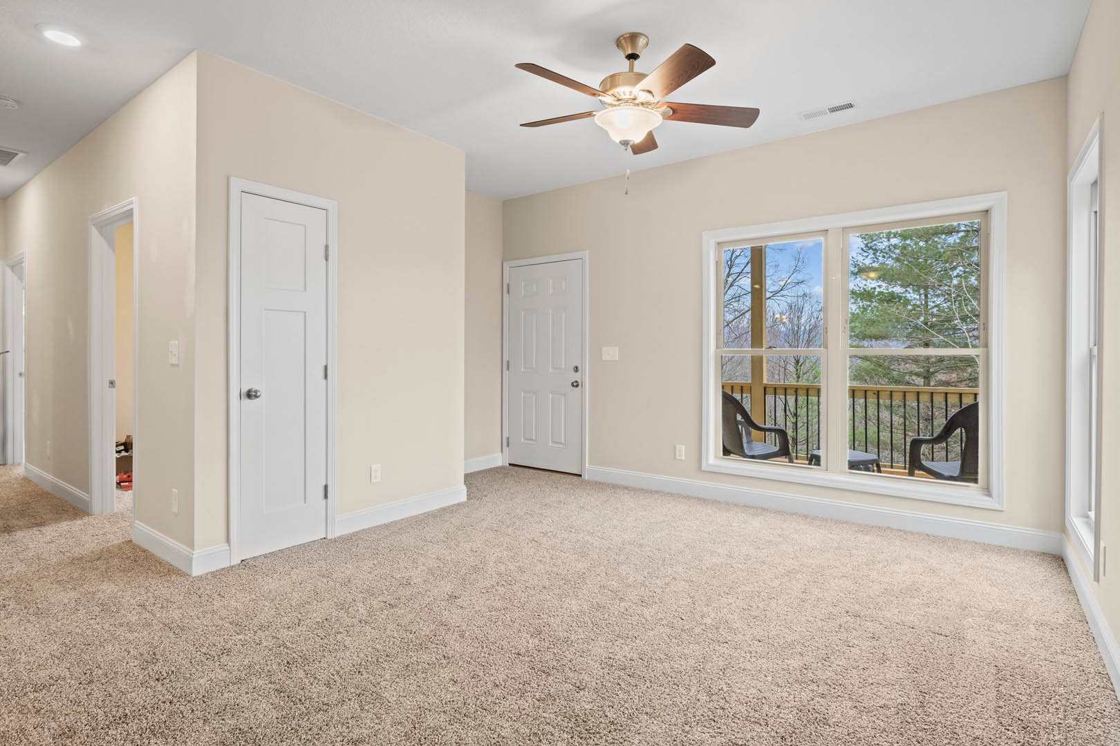 Bedroom with wood laminate flooring, ceiling fan with light fixture, white door with silver knob, large window opening to balcony with railing, outdoor table and chairs visible.