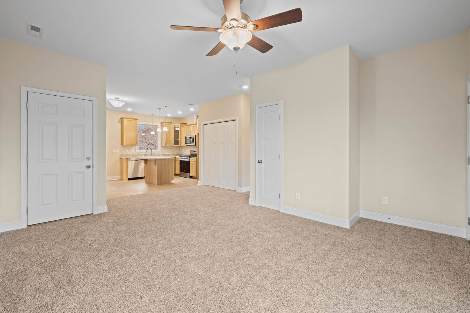 Open living room with beige carpet, white doors with silver handles, ceiling fan with light fixture, and adjacent kitchen area visible in background.
