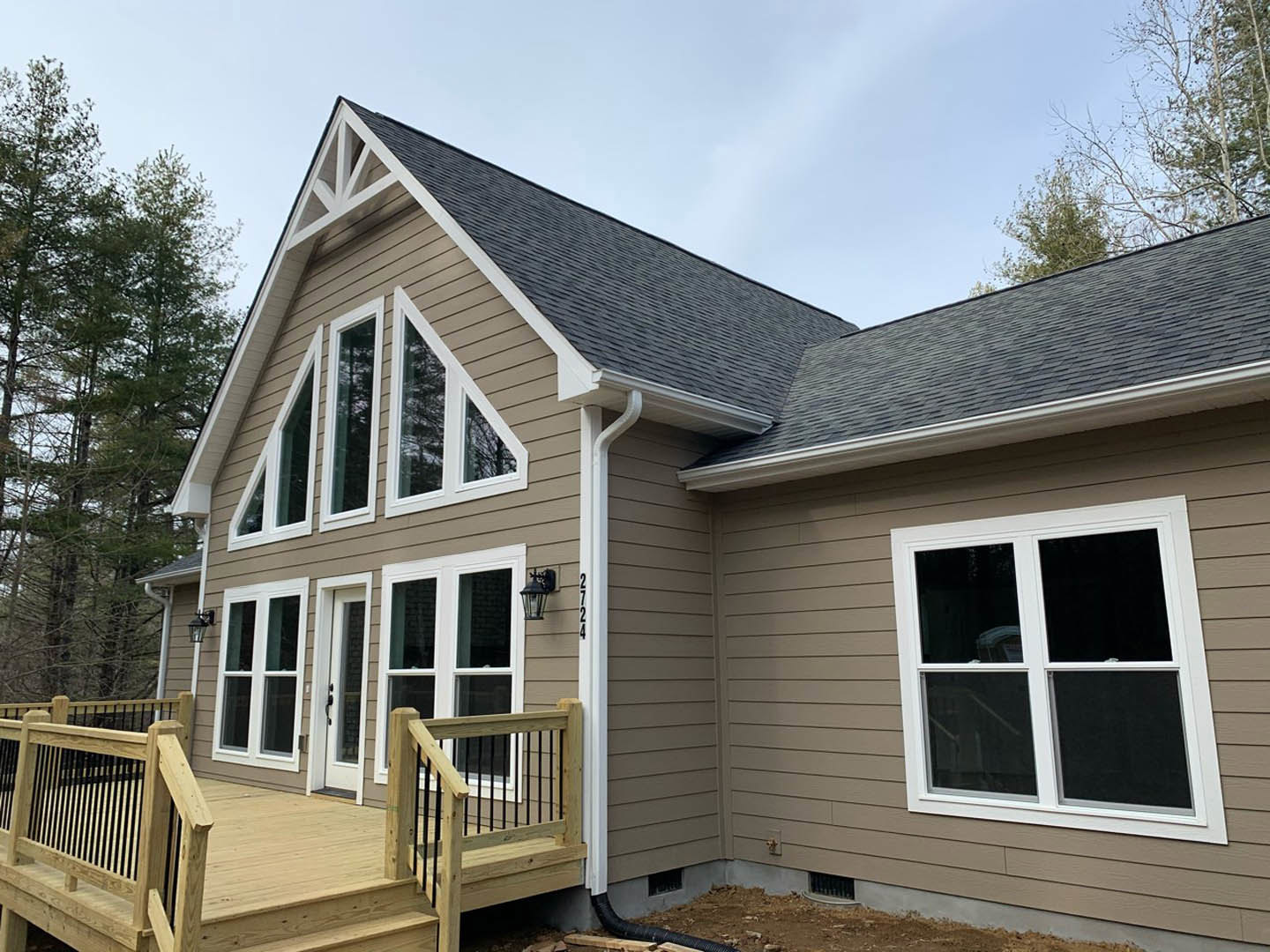 Wooden deck with railings and stairs attached to a house with light-colored siding, large windows, and leafy tree nearby