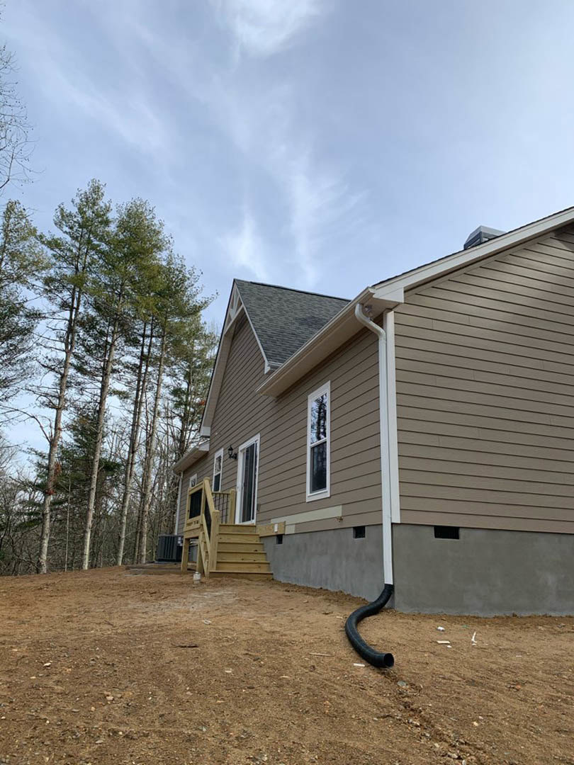 Modern wood-sided home with exterior staircase, black pipe along the ground, surrounded by tall trees under a partly cloudy blue sky