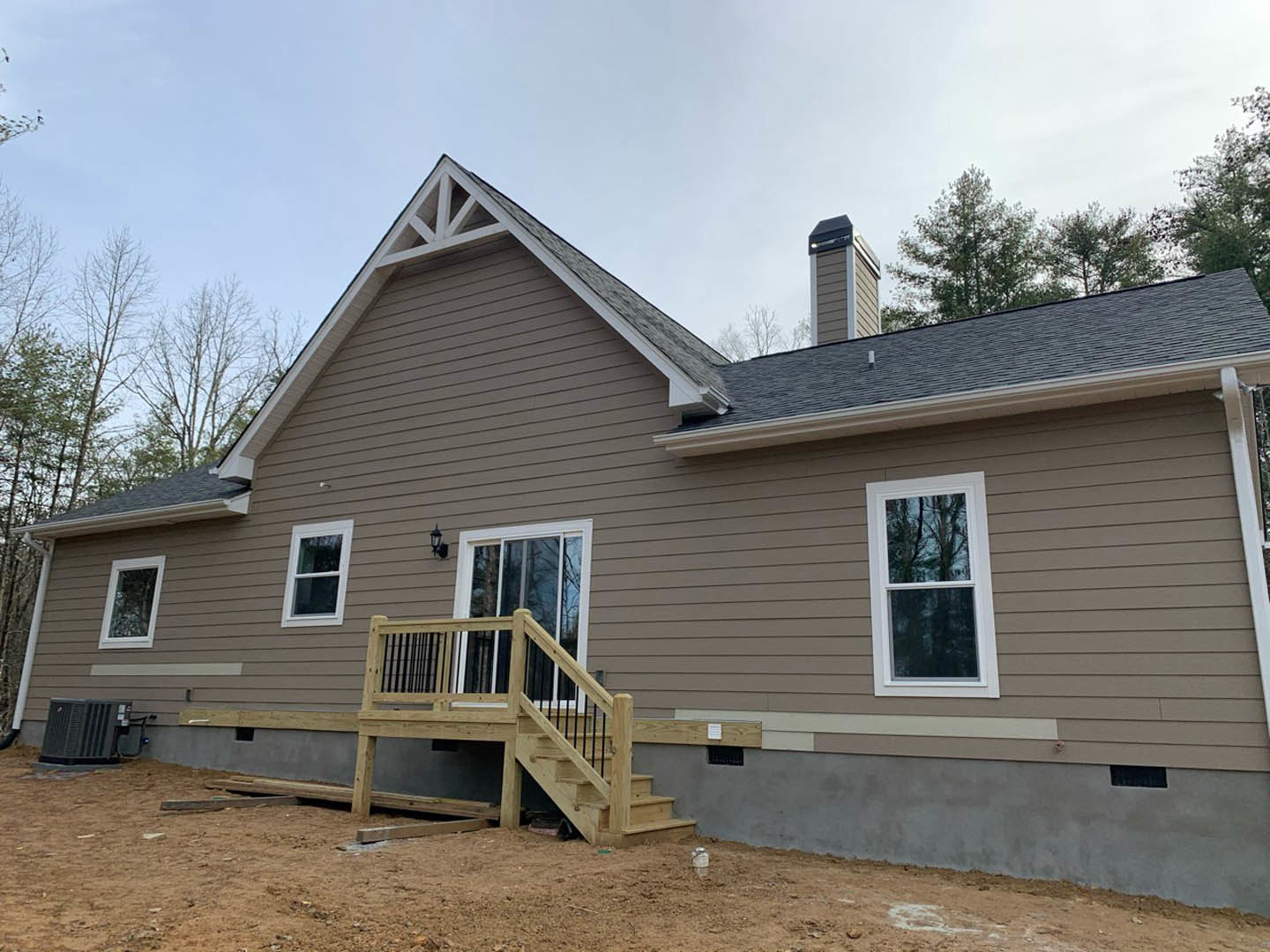 Two-story home with light siding, covered porch, wooden stairs and railing, large window reflecting trees, black air conditioning unit on ground