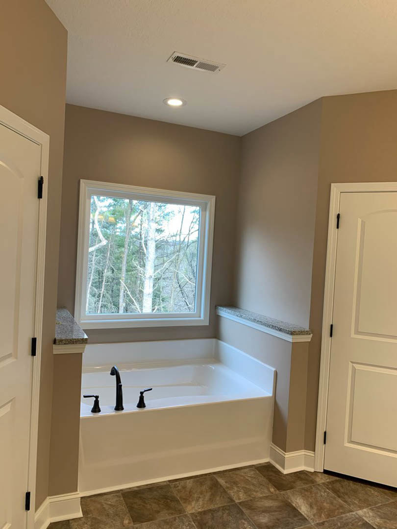 Freestanding white bathtub beside large window with tree views, light tile flooring, white door and door frame, ceiling vent, and neutral cabinetry in modern bathroom
