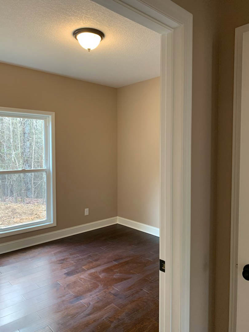 Dark hardwood floor, white walls, large window with trees visible outside, modern pendant light fixture, white door frame with brown door