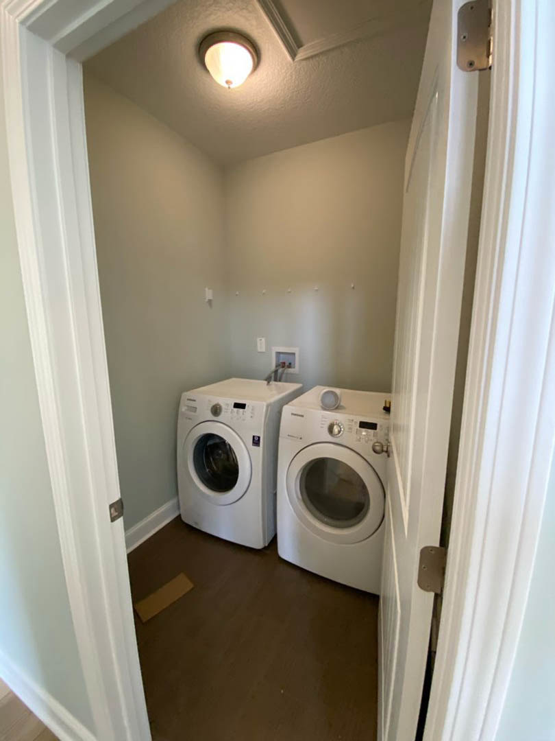 Front-loading washing machine and dryer set against white cabinetry in a laundry room with light gray walls, overhead ceiling light, and dark countertop.
