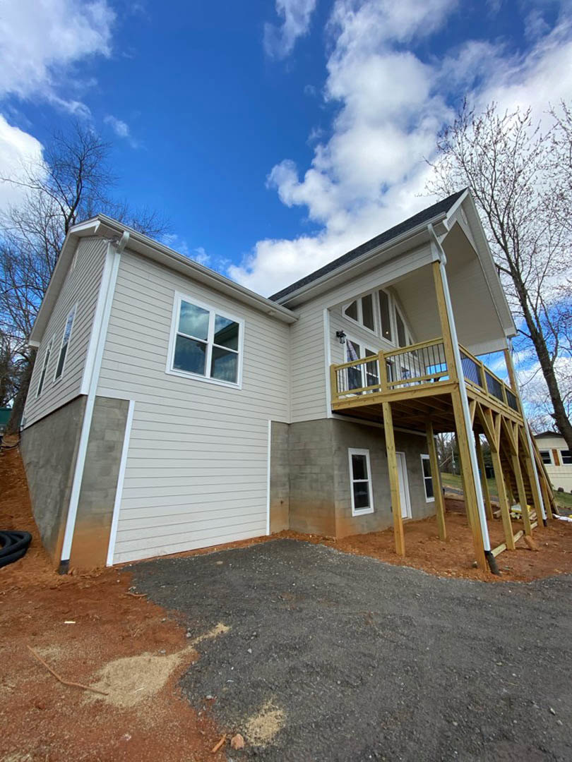 Two-story home with light siding, elevated wooden deck, gravel driveway, large windows, and mature tree in front yard