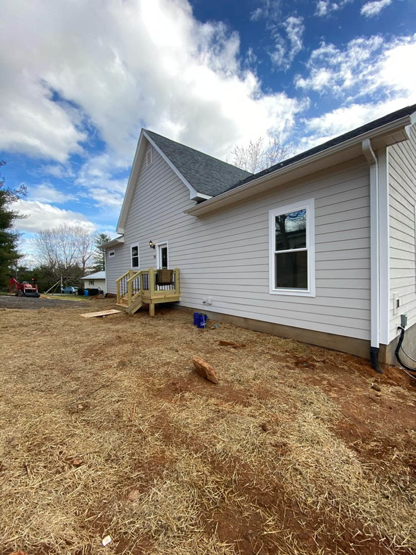 Two-story home with light siding, large windows reflecting nearby trees, wooden deck with a single chair, grassy yard bordered by rocks, under a partly cloudy blue sky