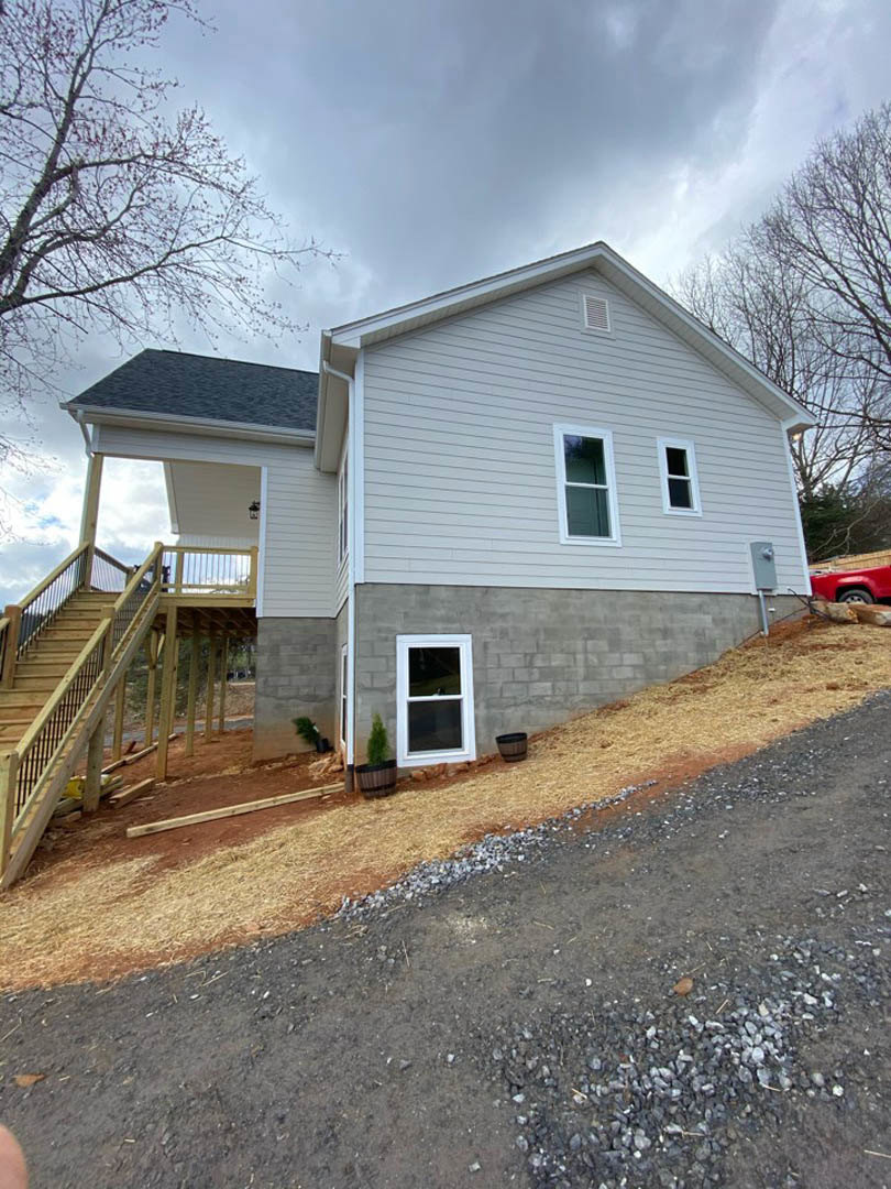 Two-story home with white-framed windows, wooden deck, and metal-railed staircase on a steep gravel slope; red truck parked beside the house; trees and cloudy sky in background.
