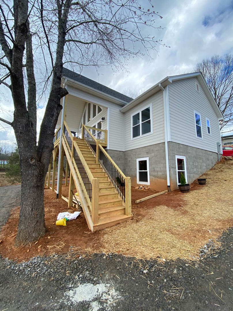 Wooden exterior staircase leading to porch, white-framed window, tree beside house, yellow bucket on ground, light-colored siding.