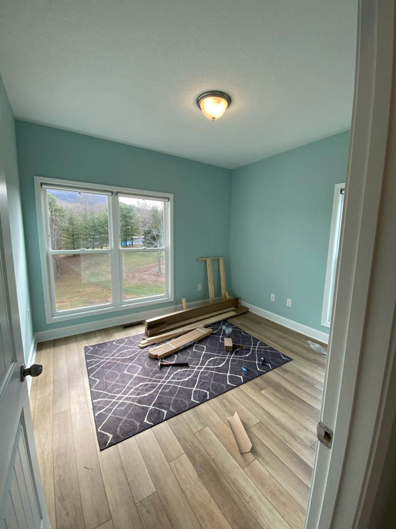Living room with wide plank wood flooring, neutral area rug, large window framing leafy trees, white plaster walls, and modern ceiling light fixture