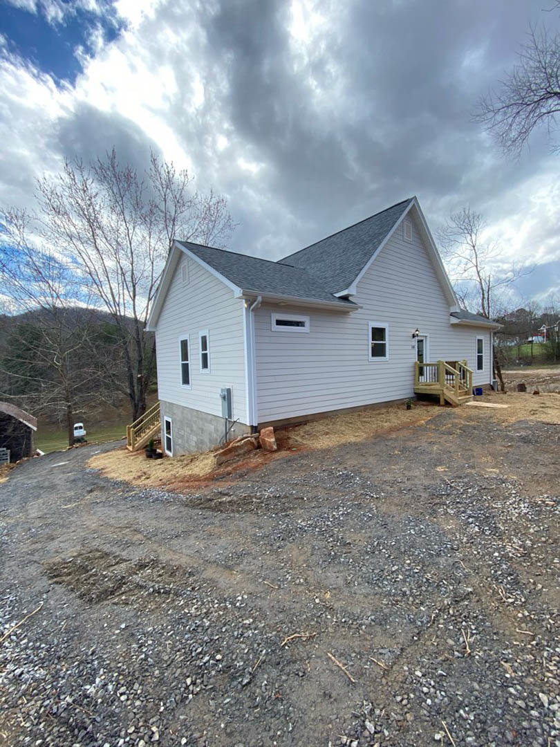 Wood-framed house under construction with exposed wooden deck and staircase, gravel driveway in foreground, surrounded by mature trees