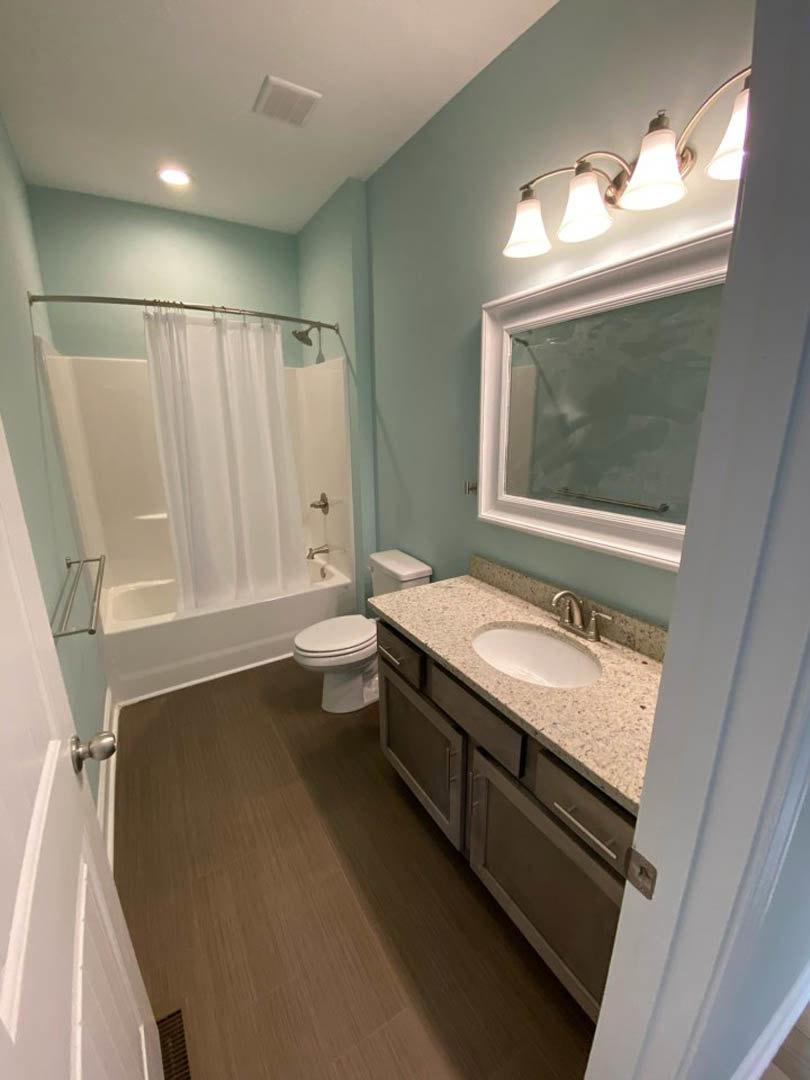 Modern bathroom with white tile walls, a pedestal sink, toilet, shower with curtain, three-light fixture above mirror, and light-colored countertop.