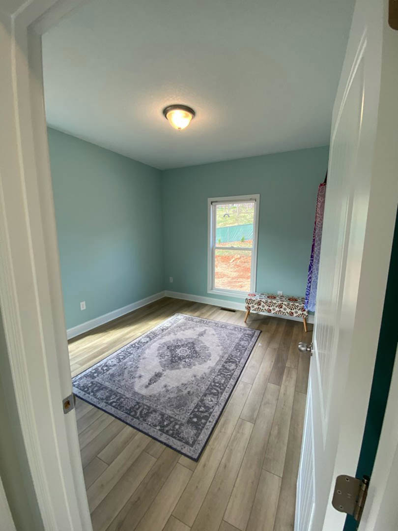 Bedroom with laminate wood flooring, floral-patterned bedspread, area rug, plaster walls, ceiling light fixture, and large window overlooking a fenced yard.