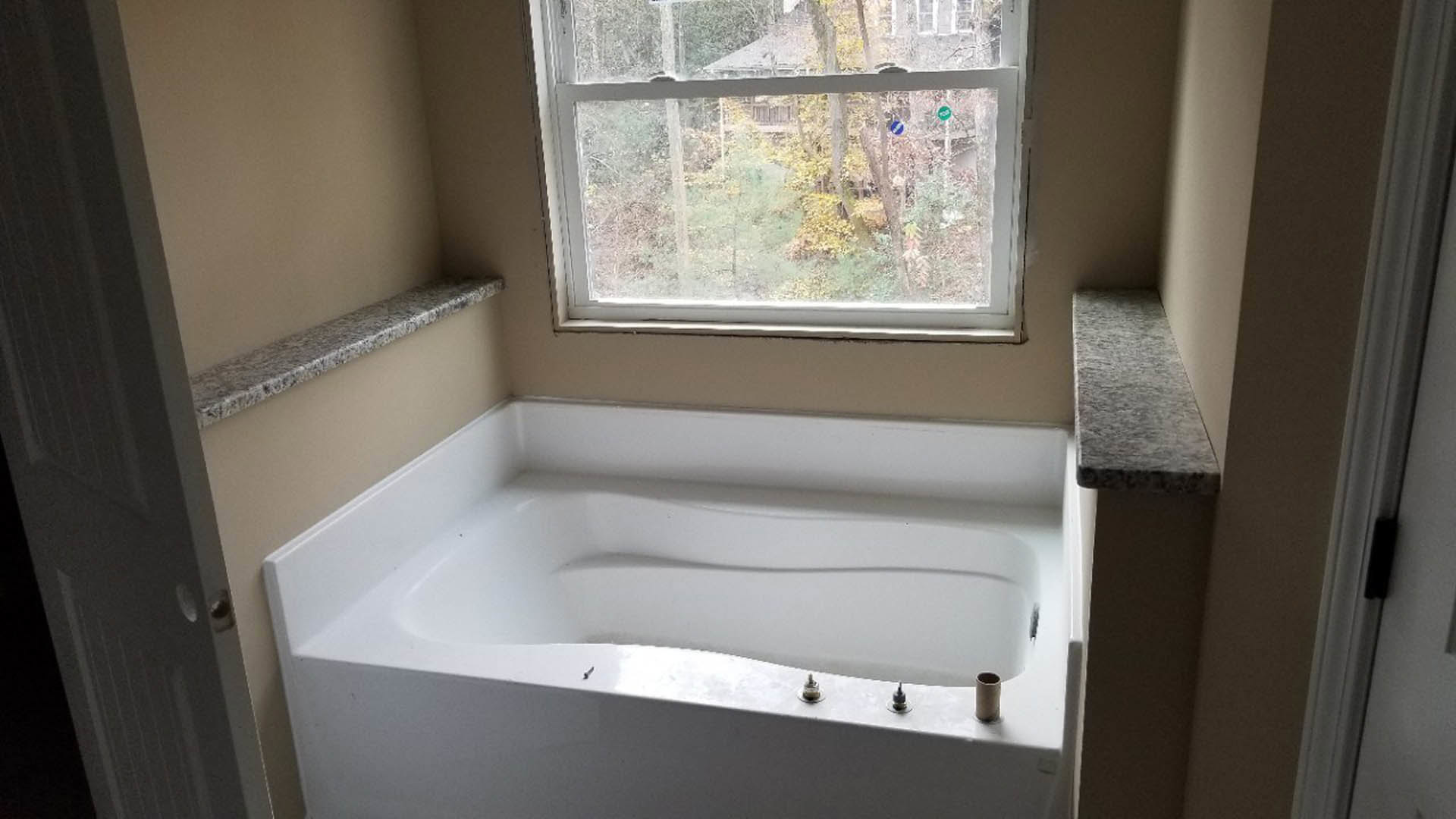 Freestanding white bathtub with corner drain beside large window overlooking trees and neighboring house, marble countertop and shelf, plaster walls, modern black faucet