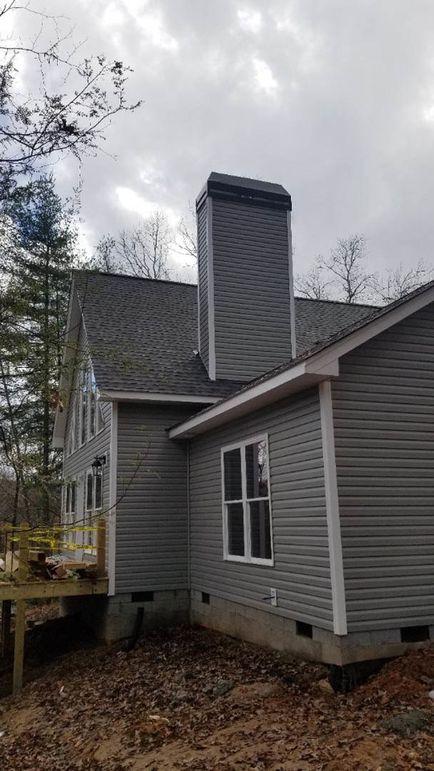 Two-story cottage with grey siding, brick chimney, covered porch, wooden deck, large windows, and mature trees in the yard.