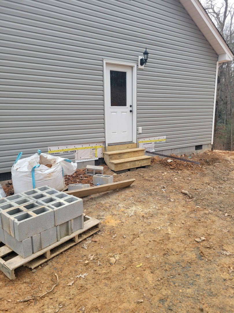 Brick exterior under construction with white door featuring glass window, stacked grey blocks on pallet, large white bag with blue straps, wooden stairs on pallet, outdoor light