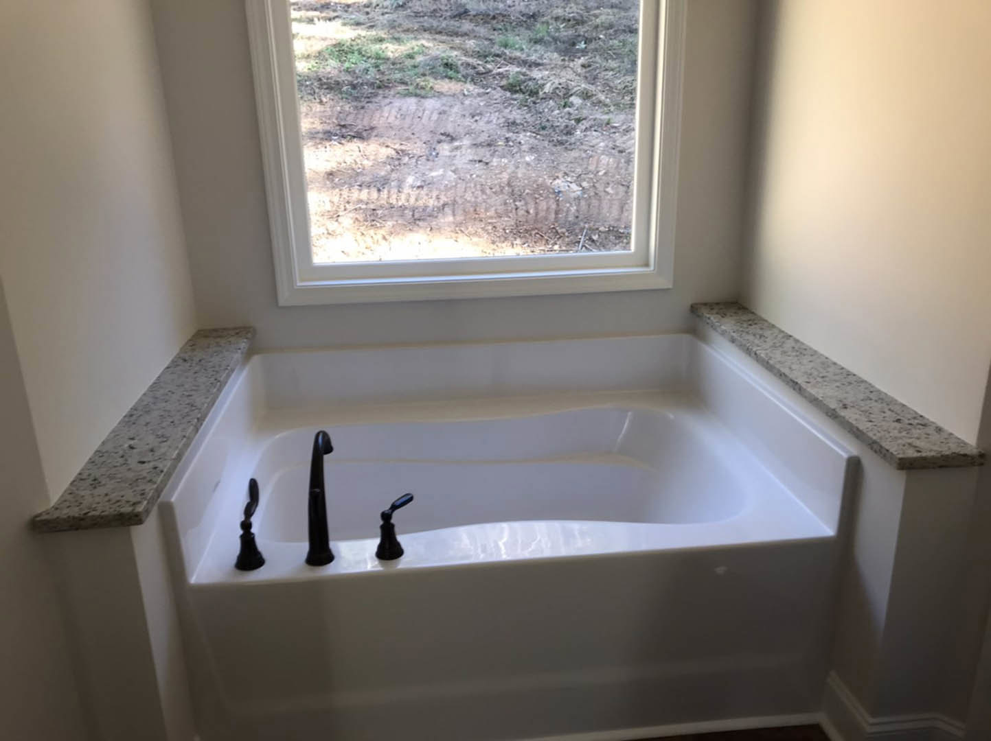 White freestanding bathtub with matte black faucets beneath a large window overlooking a dirt field, adjacent to a light-colored countertop in a modern bathroom.