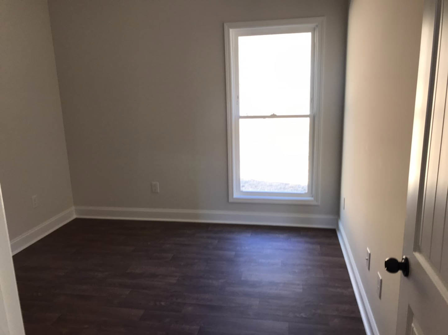 Bright room with white-framed window, dark wood flooring, white baseboards, and a close-up of a brushed metal door knob