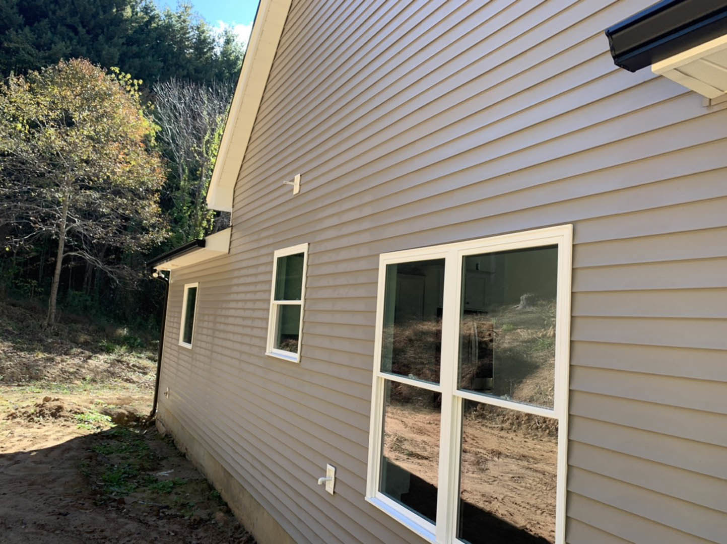Modern home exterior with light-colored siding, large window reflecting dirt field, leafless tree nearby, and clear sky in background