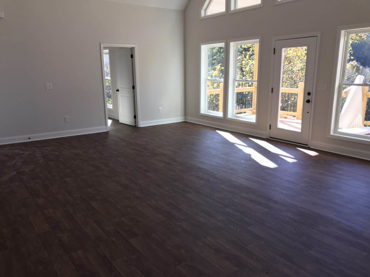 Wood floor room with large windows, white door featuring glass panel, sunlight streaming onto pale floor, view of trees and railing outside