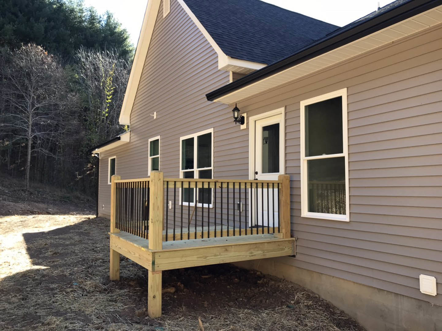 Two-story home with gray siding, large windows, and wooden deck featuring metal railing; mature trees and blue sky in background