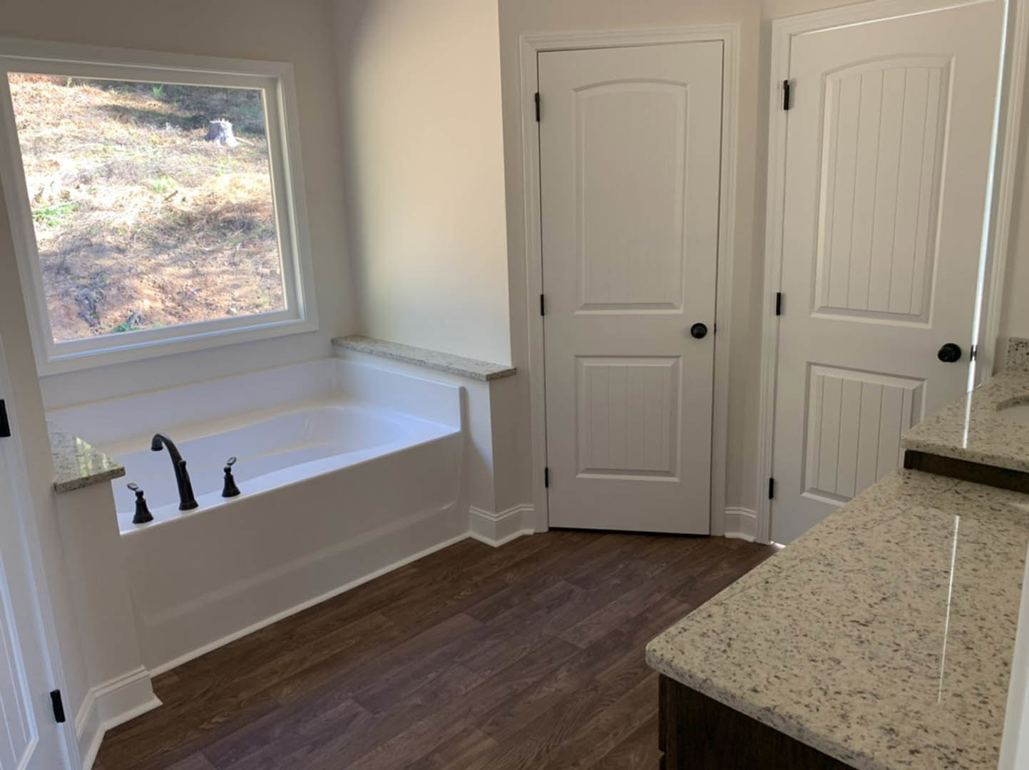 Bathroom featuring a freestanding bathtub, white speckled countertop sink, wood flooring, white door with black knobs, and window overlooking a grassy field.